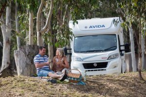 Couple enjoying a picnic in front of an Avida motorhome surrounded by trees, illustrating the lifestyle and adventure of owning a motorhome on the Gold Coast.