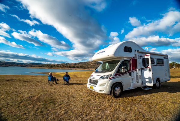 Avida motorhome parked by a serene lake, with two people relaxing in chairs, showcasing outdoor adventure and comfort in motorhome living.