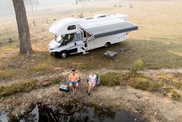Avida motorhome parked by a serene waterway, with two individuals relaxing nearby, enjoying the outdoor lifestyle and freedom of travel.