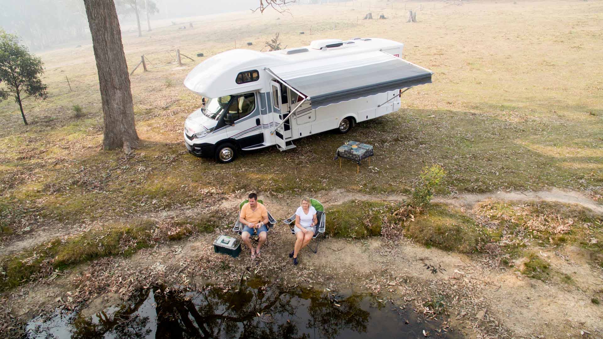 Couple relaxing beside Avida motorhome in natural setting, showcasing outdoor lifestyle and travel comfort.
