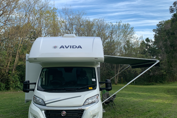 Avida motorhome parked in a grassy area with an extended awning, showcasing the brand logo on the front and surrounded by trees.