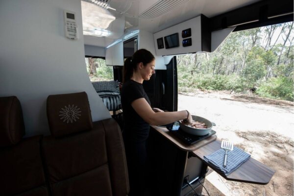 Woman cooking on a portable induction cooktop inside the Avida Explorer LX 4&times;4 campervan, featuring leather seating and a view of a natural outdoor setting.