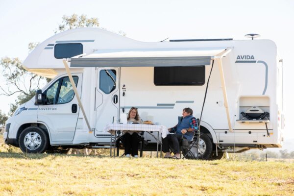Couple enjoying outdoor dining under the electric awning of the Avida Silverton motorhome, showcasing its practical design and outdoor living features.