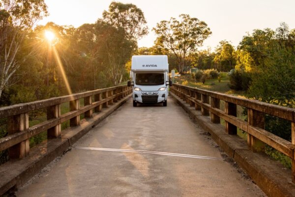Avida Silverton motorhome driving on a bridge at sunset, surrounded by trees, showcasing outdoor adventure and travel lifestyle.