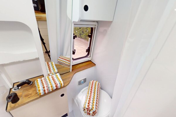 Compact campervan bathroom with modern fixtures, mirror, and storage, featuring a toilet and colorful towels, designed for the Avida Trek campervan.