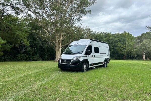 Avida Trek campervan parked on grassy field, showcasing sleek design and compact size, surrounded by trees and cloudy sky, emphasizing suitability for adventurous travel.