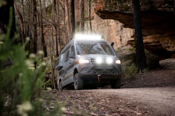 Avida Explorer LX 4&times;4 campervan driving on a forest trail, showcasing off-grid features with rooftop lights and rugged design, surrounded by trees and rocky terrain.