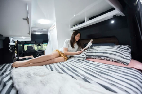Woman relaxing on striped bedding in Avida Explorer LX 4&times;4 campervan, reading a book, showcasing the camper's interior comfort and design features.