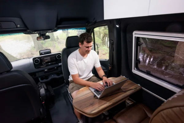 Man working on a laptop at a wooden table inside the Avida Explorer LX 4x4 campervan, showcasing its interior design and off-grid capabilities.