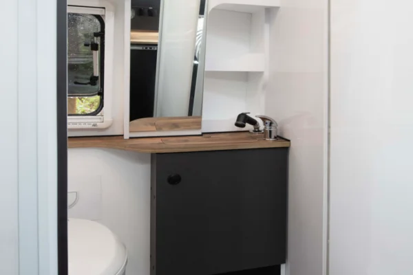 Compact campervan bathroom featuring a toilet, wooden countertop with a sink and faucet, mirror, and storage shelves, showcasing modern design elements for the Avida Explorer LX 4x4 campervan.